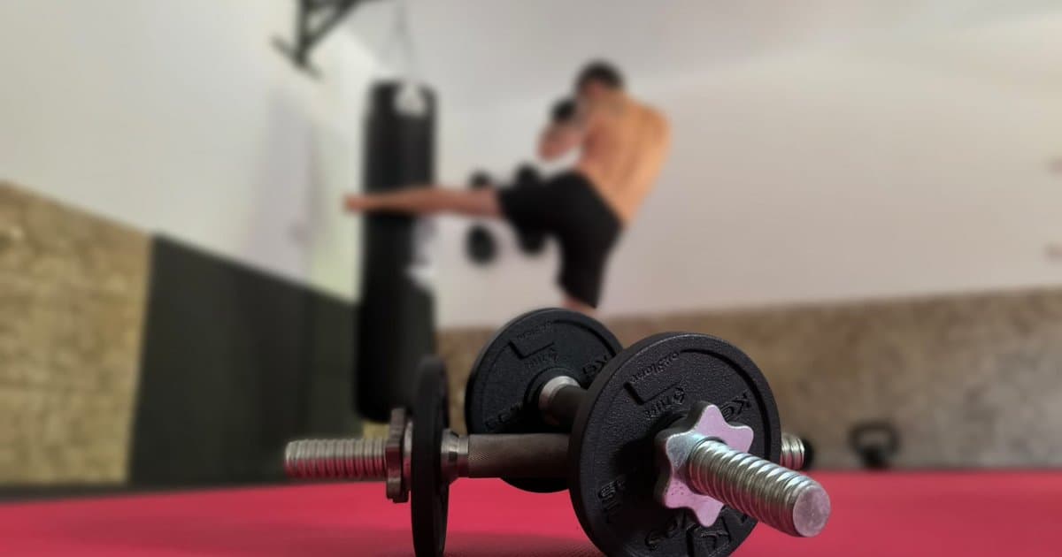 Una mancuerna con discos en primer plano sobre una esterilla roja, con un baterista entrenando con un saco de boxeo al fondo en un gimnasio