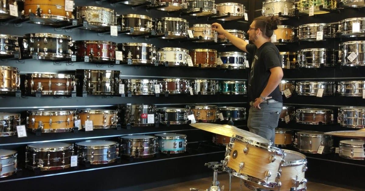 Baterista en una tienda de música rodeado de una pared llena de cajas de batería, alcanzando una de ellas, con una batería acústica de madera en primer plano