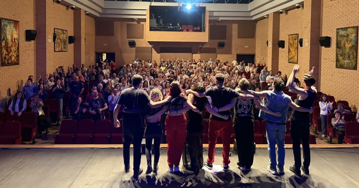 La banda saluda abrazada por los hombros al final del concierto sobre el escenario de un teatro, con el público en pie aplaudiendo