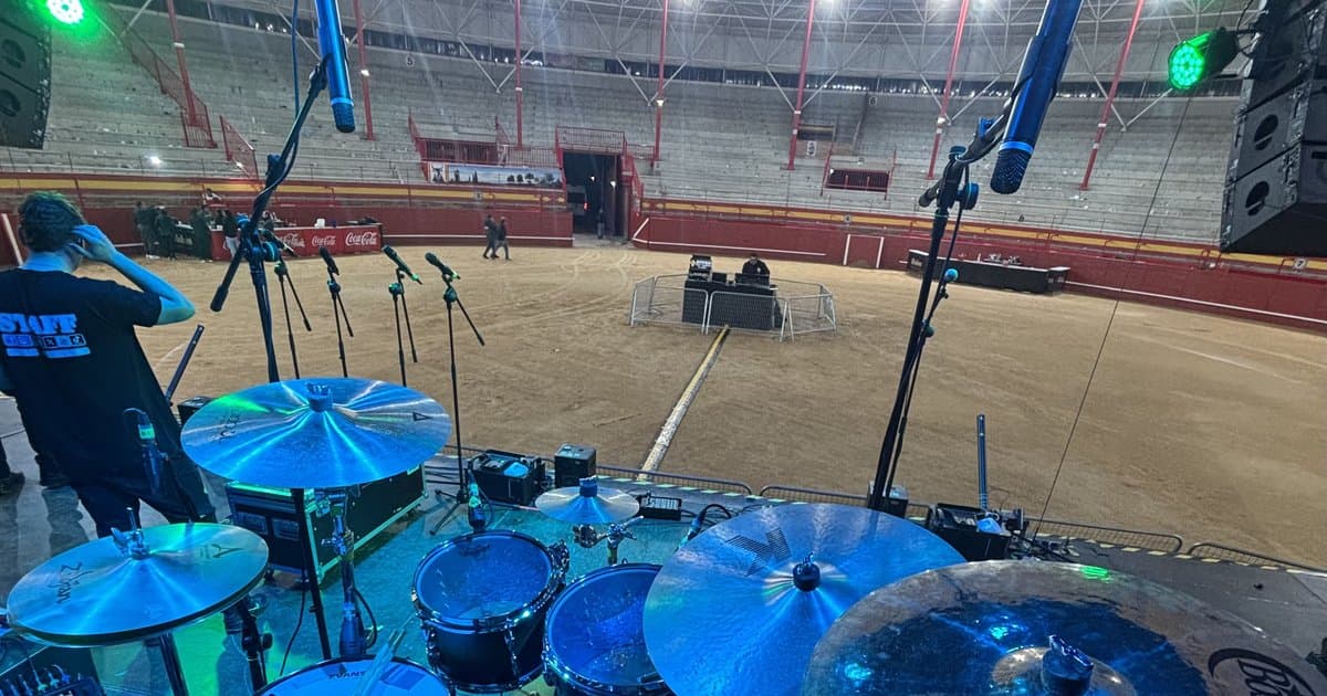 Vista desde detrás de una batería con platos azulados hacia una plaza de toros vacía con los micrófonos montados antes de un concierto