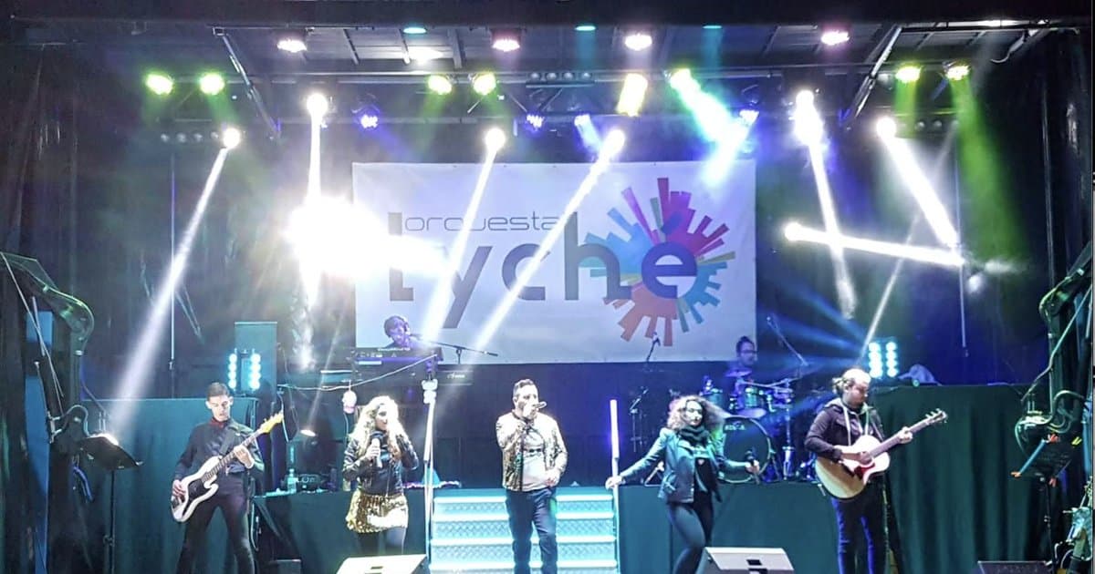 Una orquesta de baile tocando en directo sobre un escenario iluminado con focos de colores, con cantantes, guitarras y batería al fondo